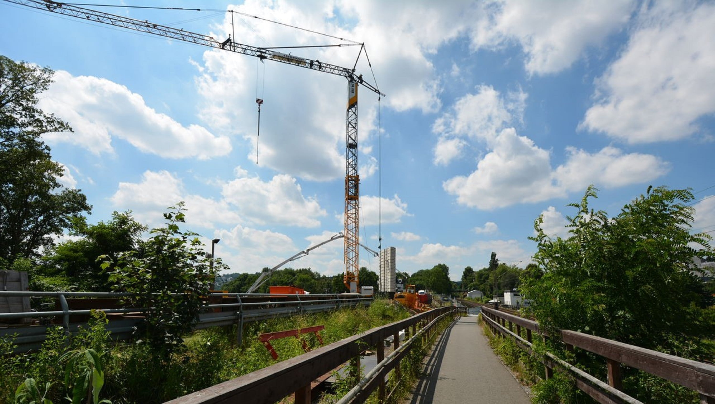 Ein hoher Baukran ragt in den blauen Himmel. Links sind Bäume und Büsche, rechts ein Fußweg mit Geländer. Baustellenfahrzeuge und Materialien sind entlang der Straße sichtbar.